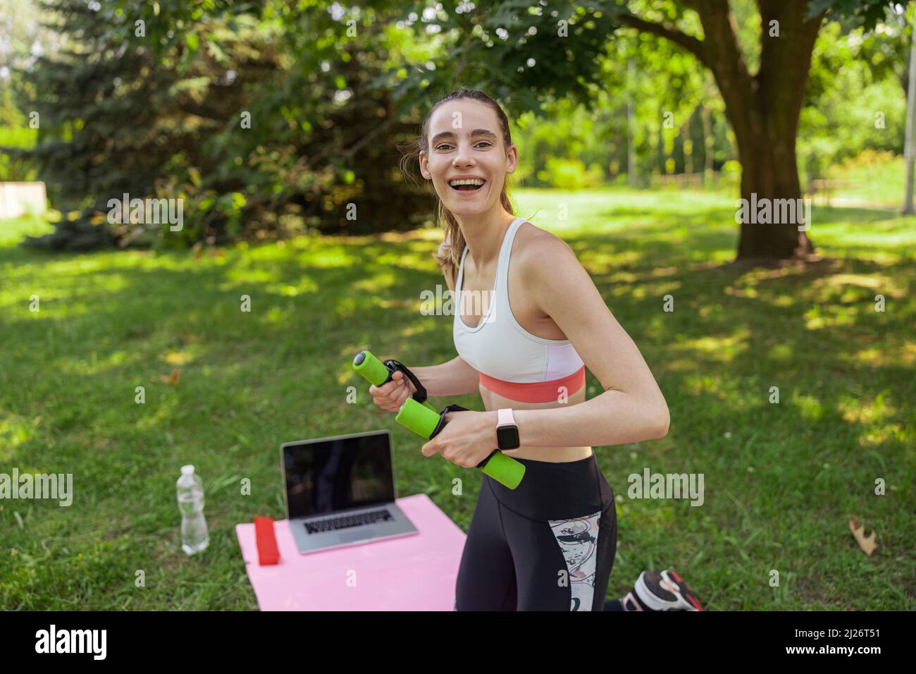 Joyful lady holds dumbbells standing on knees near laptop in green summer park Stock Photo