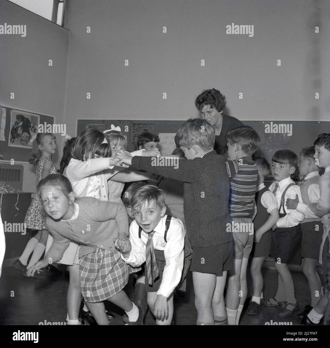 1960s, historical, primary school children playing a singing game in the classroom, possibly the one called 'Oranges and Lemons', where children move or dance through an arch made by children holding hands, facing each other, arms raised, Fife, Scotland, UK. The game is also known as 'London Bridge is Falling Down', where again two or more children join hands to form an arch, the bridge, where the other choldren march under. In Scotland it is also known as 'broken bridges'. Stock Photo