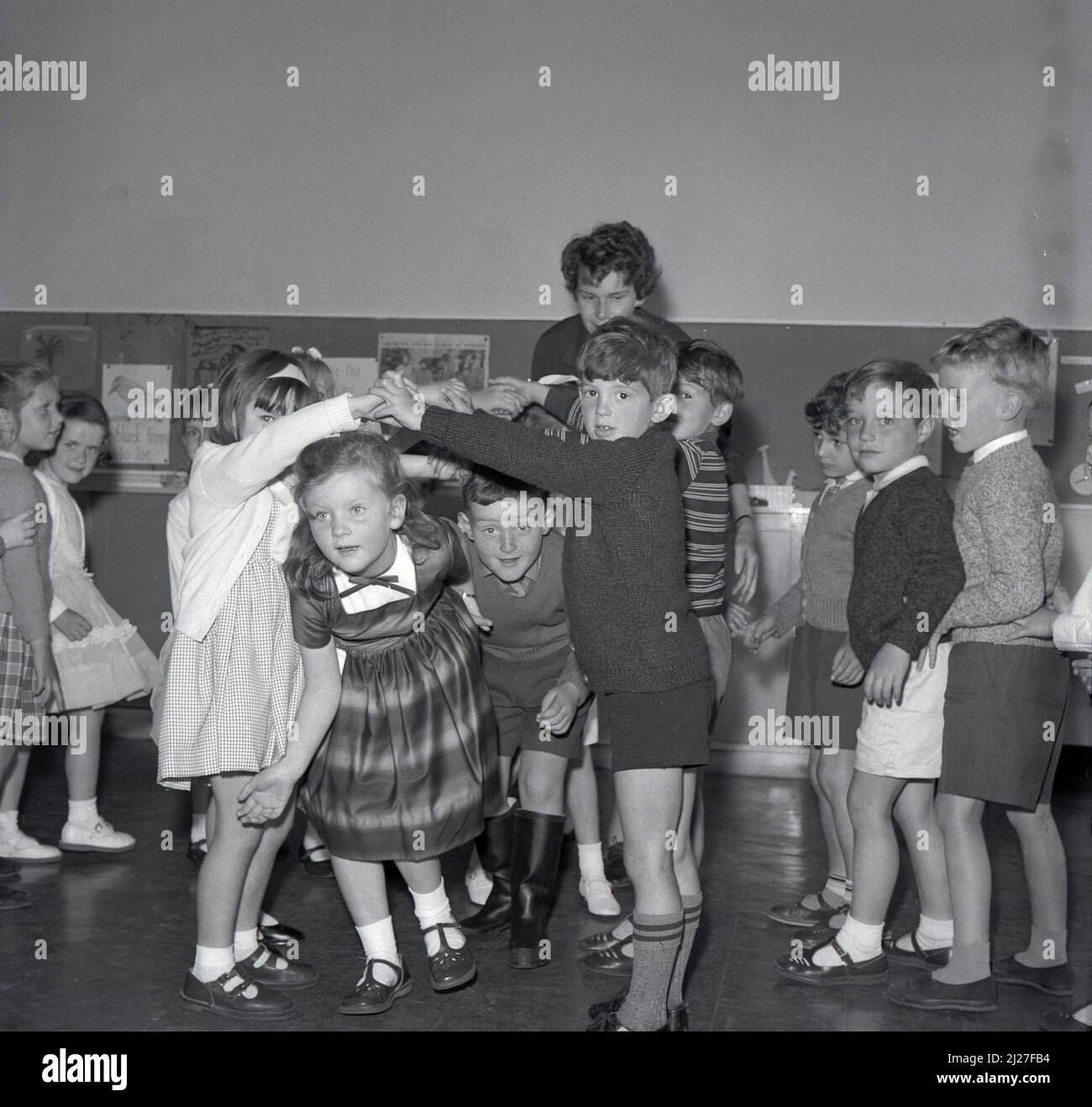 1960s, historical, primary school children playing a singing game in the classroom, possibly the one called 'Oranges and Lemons', where children move or dance through an arch made by children holding hands, facing each other, arms raised, Fife, Scotland, UK. The game is also known as 'London Bridge is Falling Down', where again two or more children join hands to form an arch, the bridge, where the other choldren march under. In Scotland it is also known as 'broken bridges'. Stock Photo