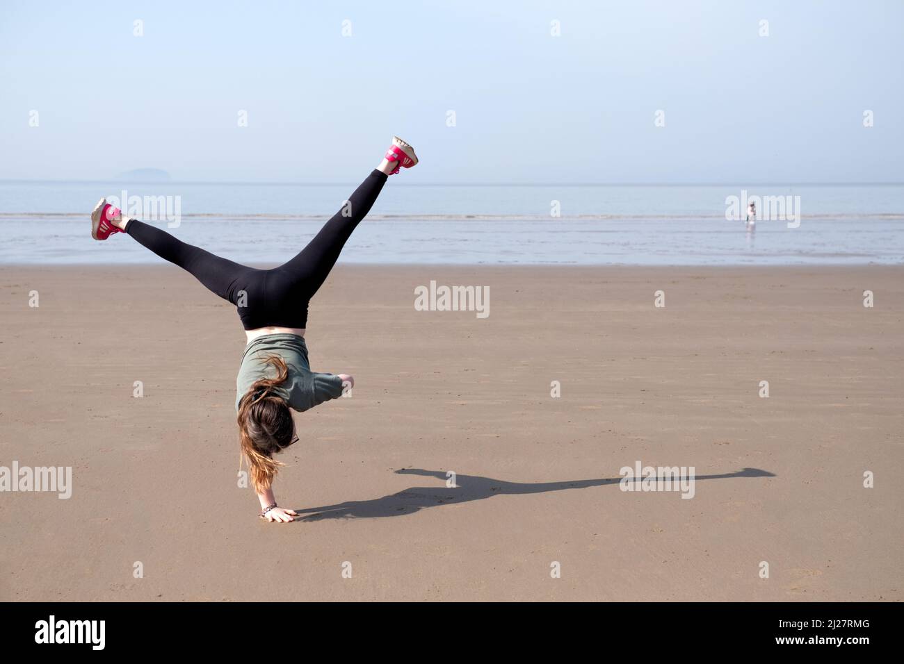 A young girl performs cartwheels on an empty beach. It’s a bright sunny day as the energetic youngster is shown mid move in an upside down position. Stock Photo