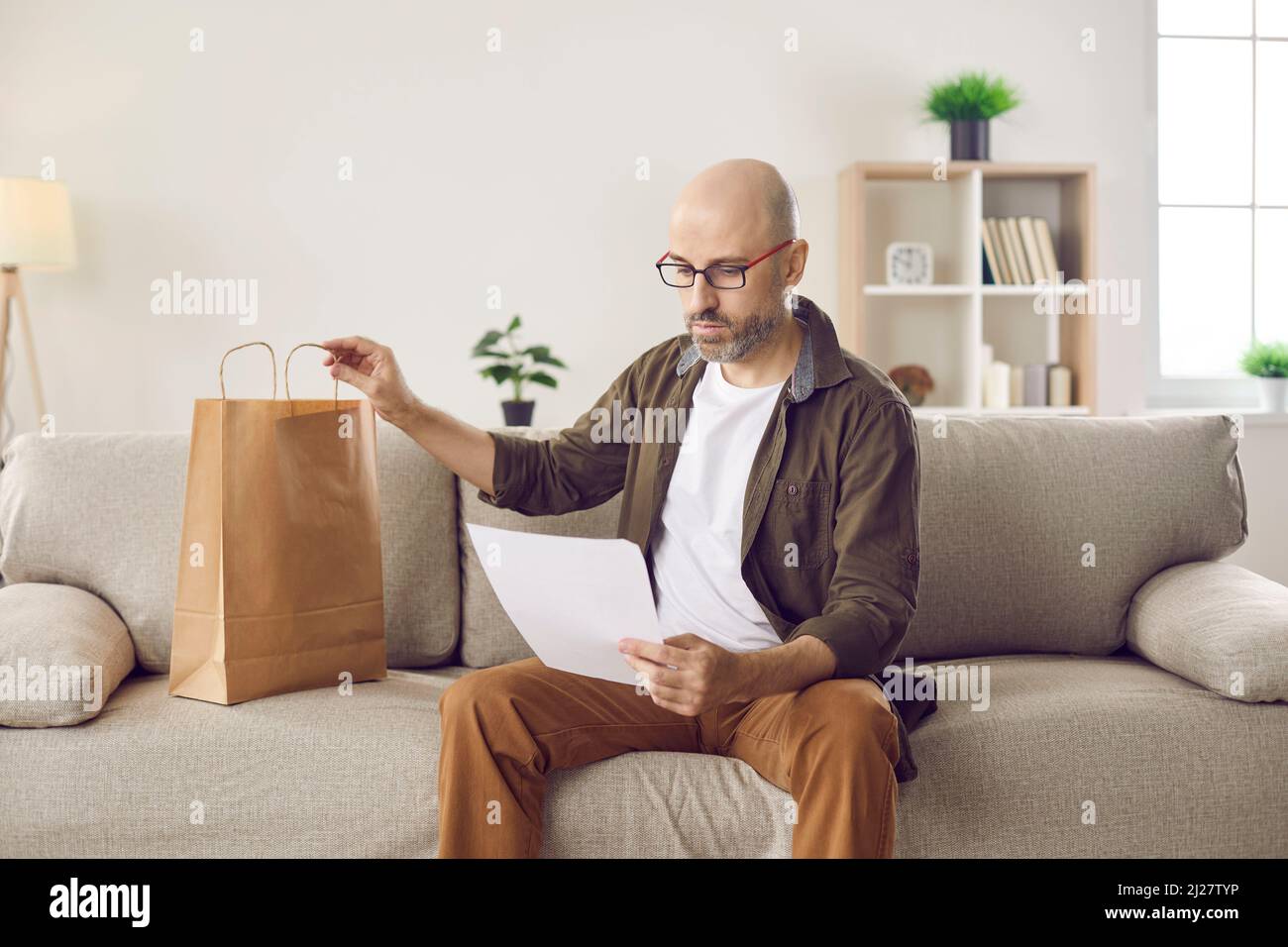 Man sitting on sofa at home and reading letter he got together with paper delivery bag Stock Photo