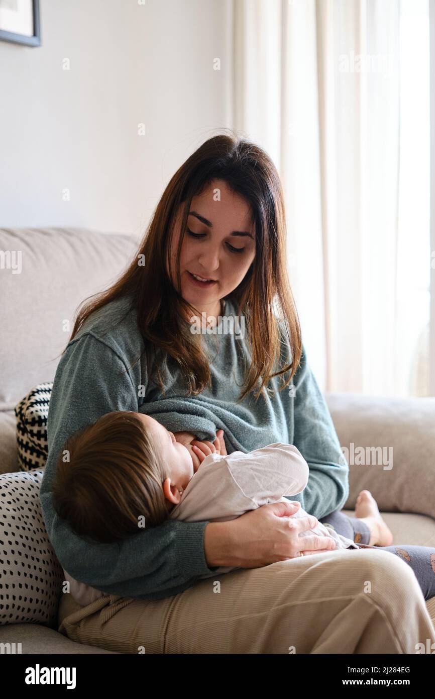 Mother breastfeeding her daughter while sitting on the sofa at home. Stock Photo