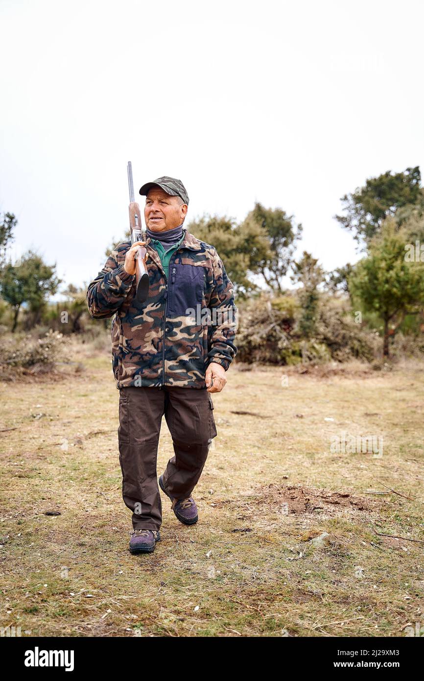 Fulllength male hunter in camouflage outerwear and cap carrying a