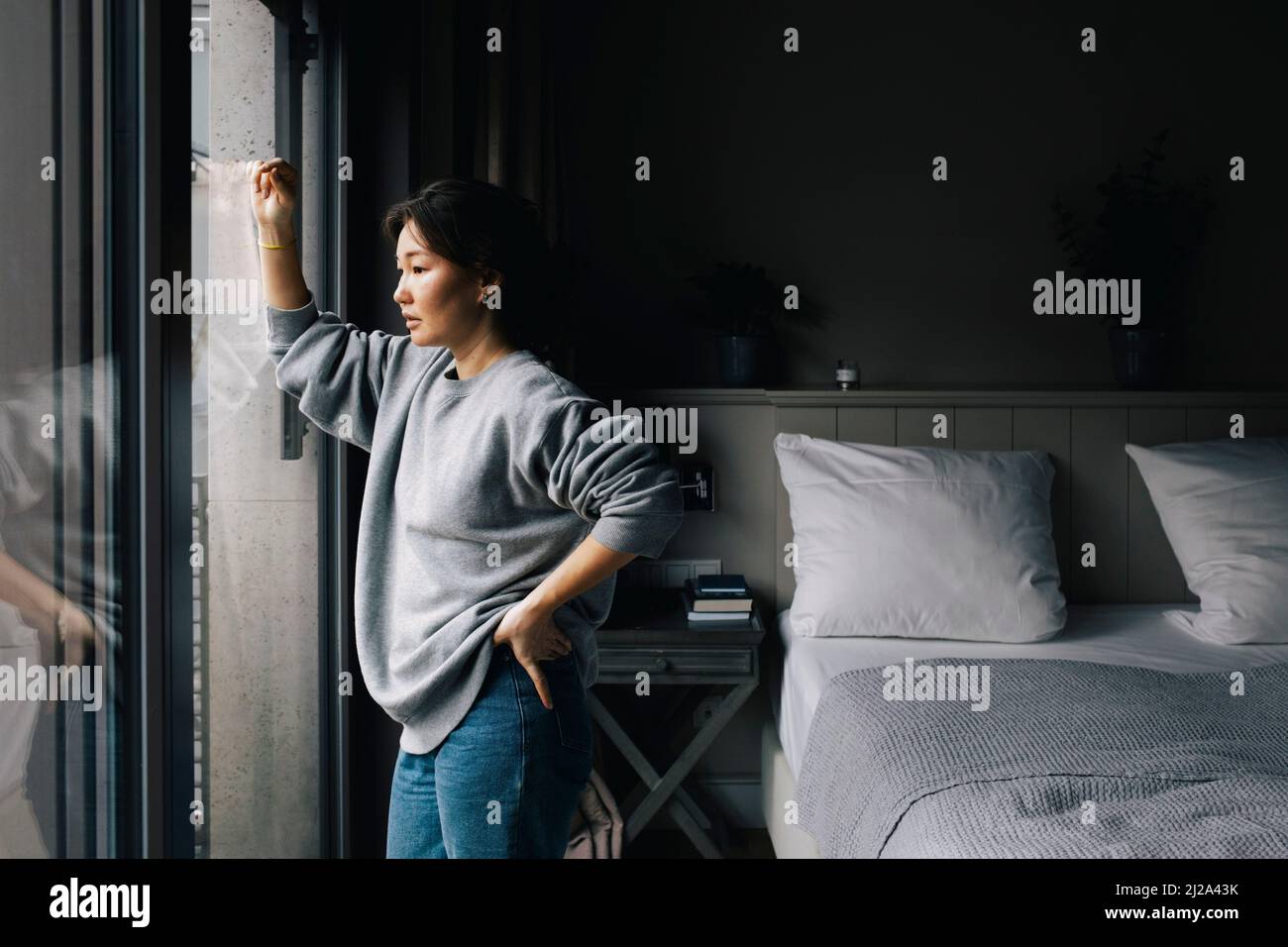 Thoughtful lonely woman looking out through window while standing with hand on hip in bedroom at home Stock Photo