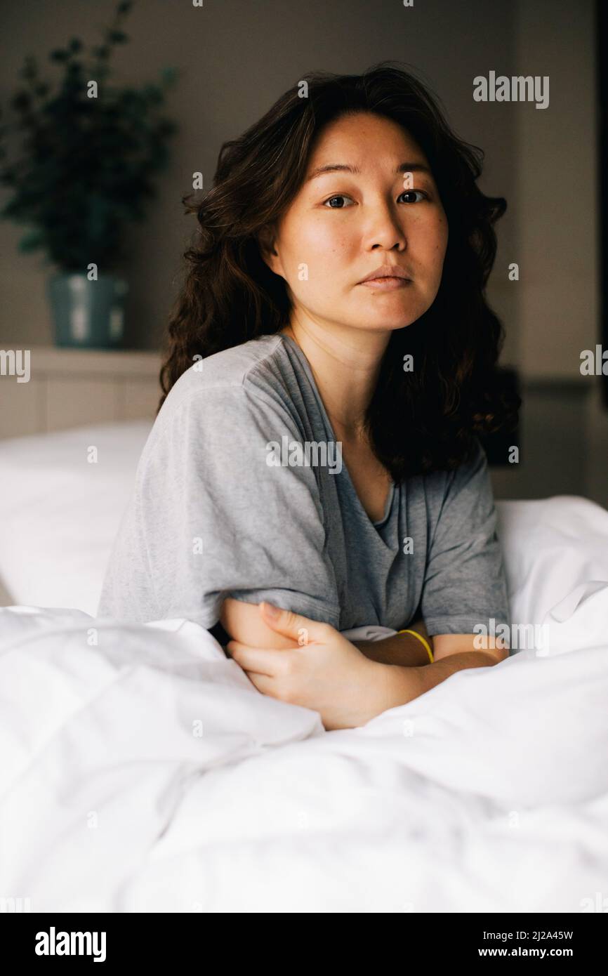 Portrait of lonely woman sitting on bed with white blanket at home Stock Photo