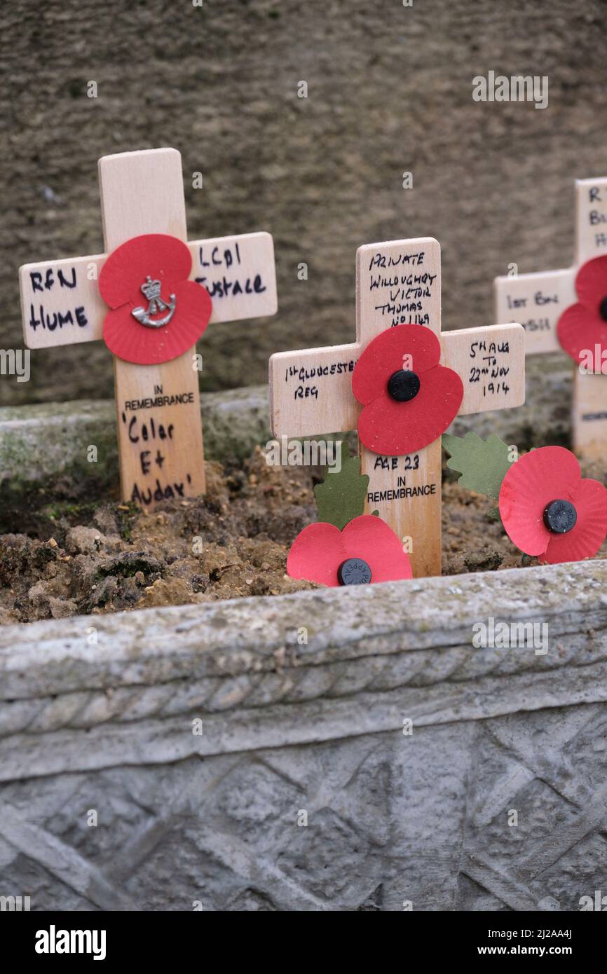 Small wodden crosses of rememberance sit against a stone memorial with faded paper poppies. Stock Photo