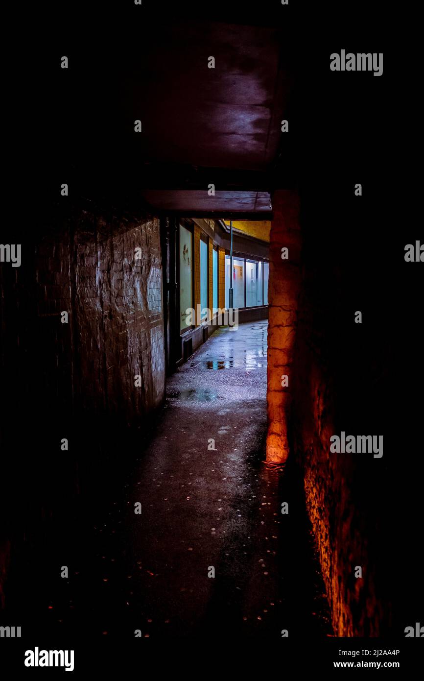 Looking through a dark alleyway at the brighlty lit shop windows in the distance on a dark wet night. Stock Photo