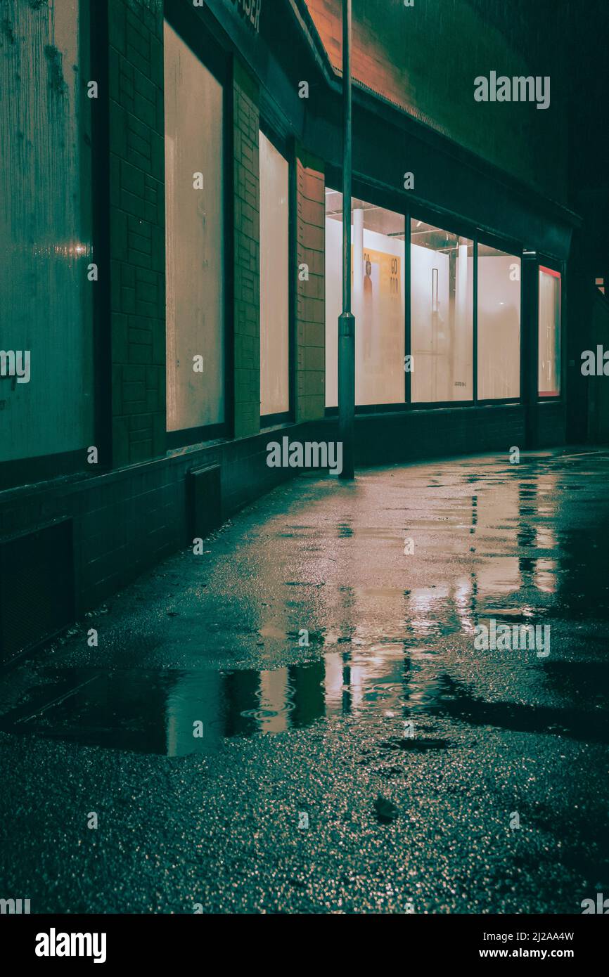 Looking through a dark alleyway at the brighlty lit shop windows in the distance on a dark wet night. Stock Photo