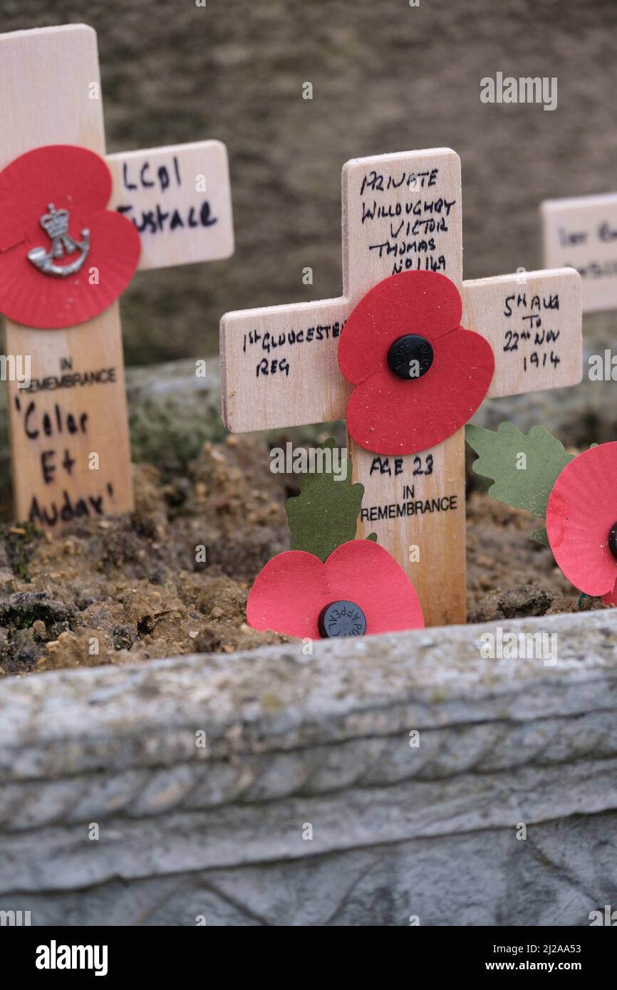 Small wodden crosses of rememberance sit against a stone memorial with faded paper poppies. Stock Photo
