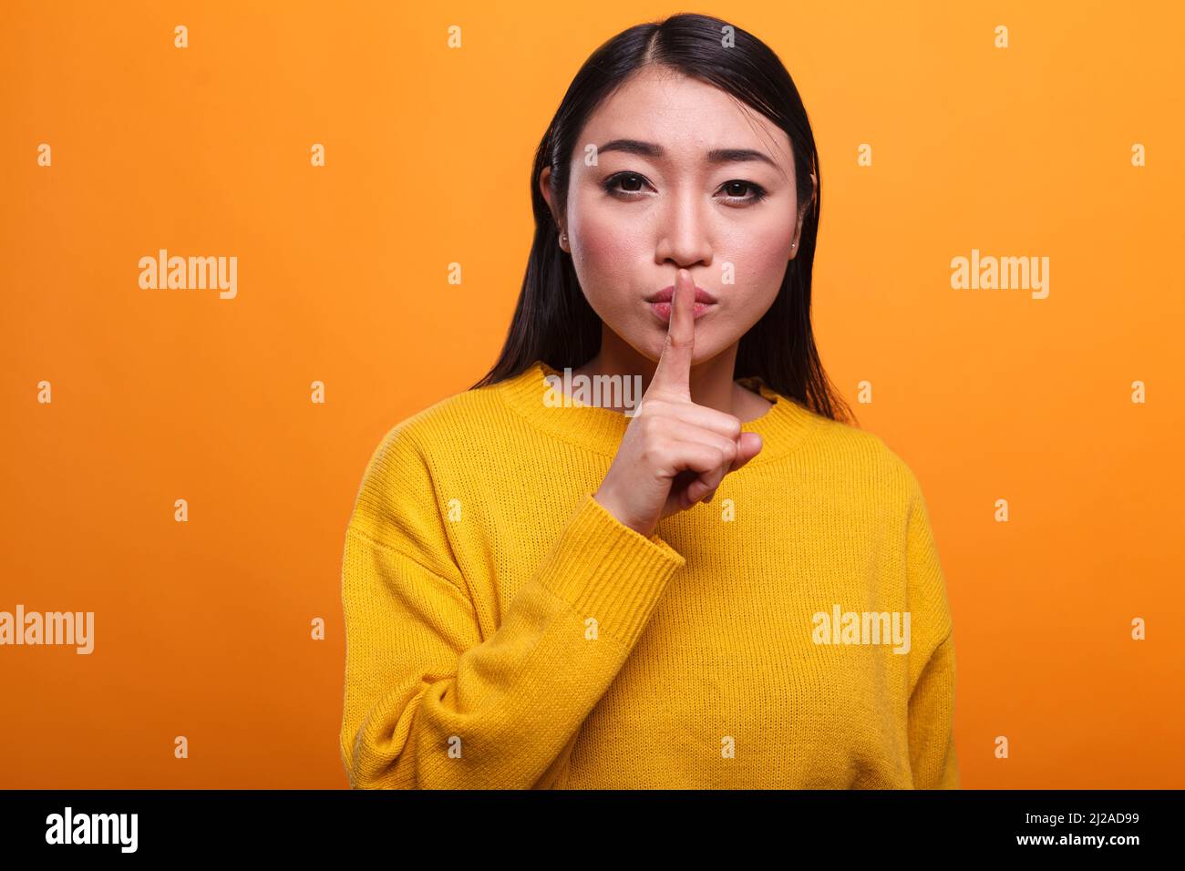 Calm attractive women wearing yellow sweater making hush gesture with forefinger on orange background. Mysterious person touching lips with index finger indicating silence and secrecy. Stock Photo