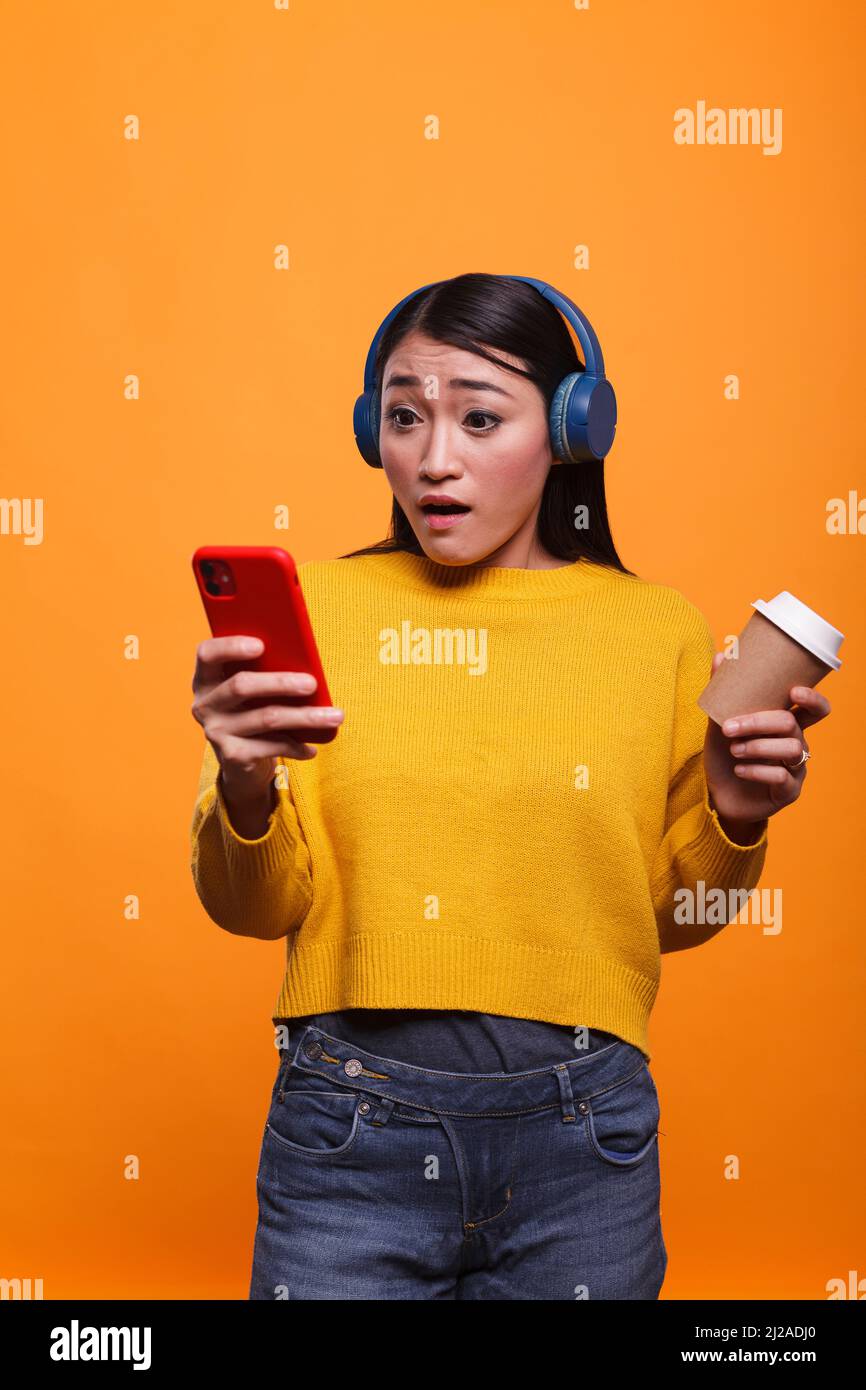 Shocked speechless young adult woman reacting in fear while reading tragic news on smartphone device. Frightened astonished asian person using mobile cellphone while left stunned. Stock Photo