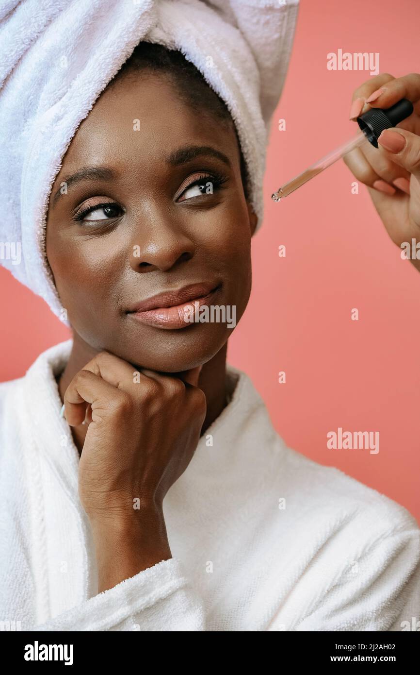 Portrait of a young woman thinking in bathrobe with spa towel on head receiving treatment with pipette on pink copy space Stock Photo
