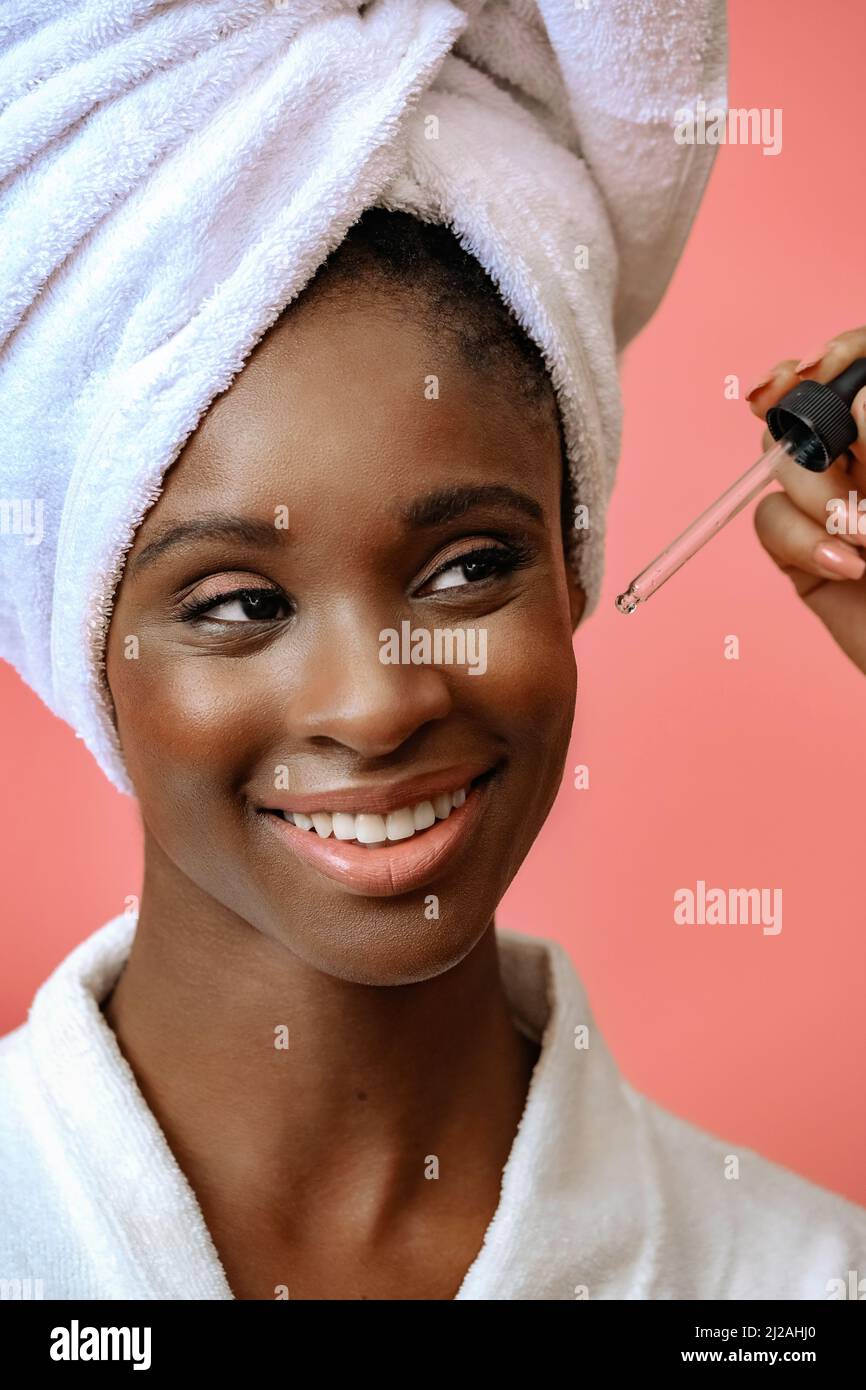 Young happy woman in bathrobe with spa towel on head receiving treatment with pipette on pink copy space Stock Photo