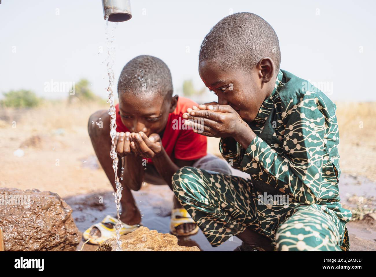 Two thirsty siblings drinking fresh water from a remote tap in the steppe of rural sub-Saharan Africa; symbol of global climate change, water scarcity Stock Photo