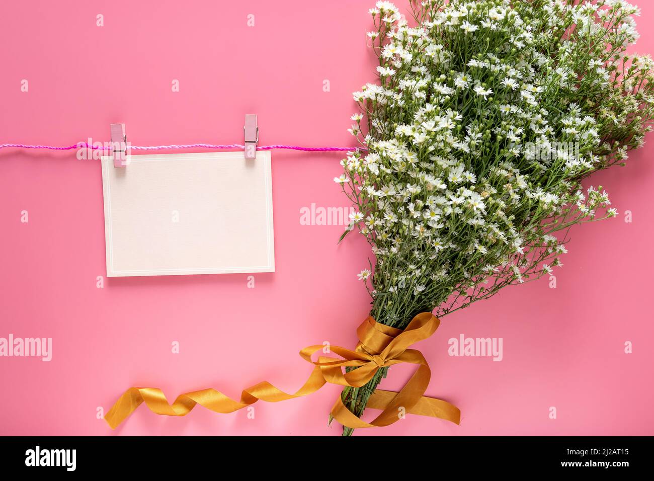 mockup of pastel pink card note blank paper sheet hang on rope with clothes peg and spring fresh white flower bouquet isolated on pink background. Stock Photo