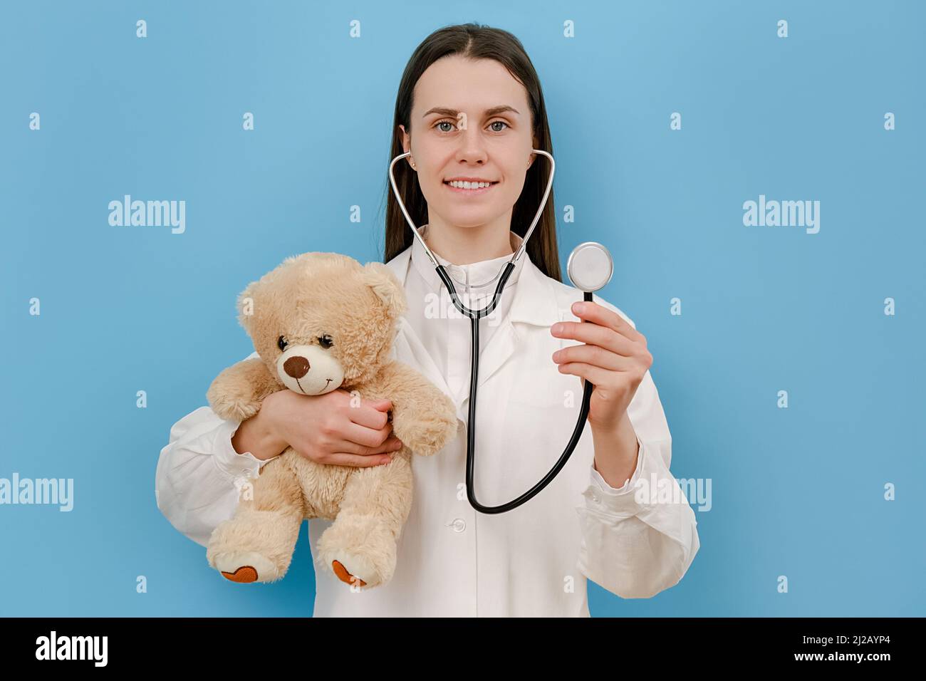 Portrait of cute smiling young woman doctor holding stethoscope and small fluffy bear, looking at camera, isolated over blue color background wall in Stock Photo