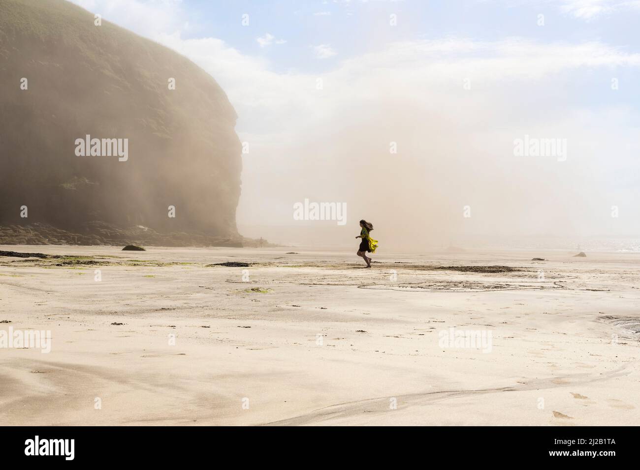 On a sunny summer day a woman runs across Druidstone beach as it is enveloped in a cloud of dust due to a rock fall. Stock Photo