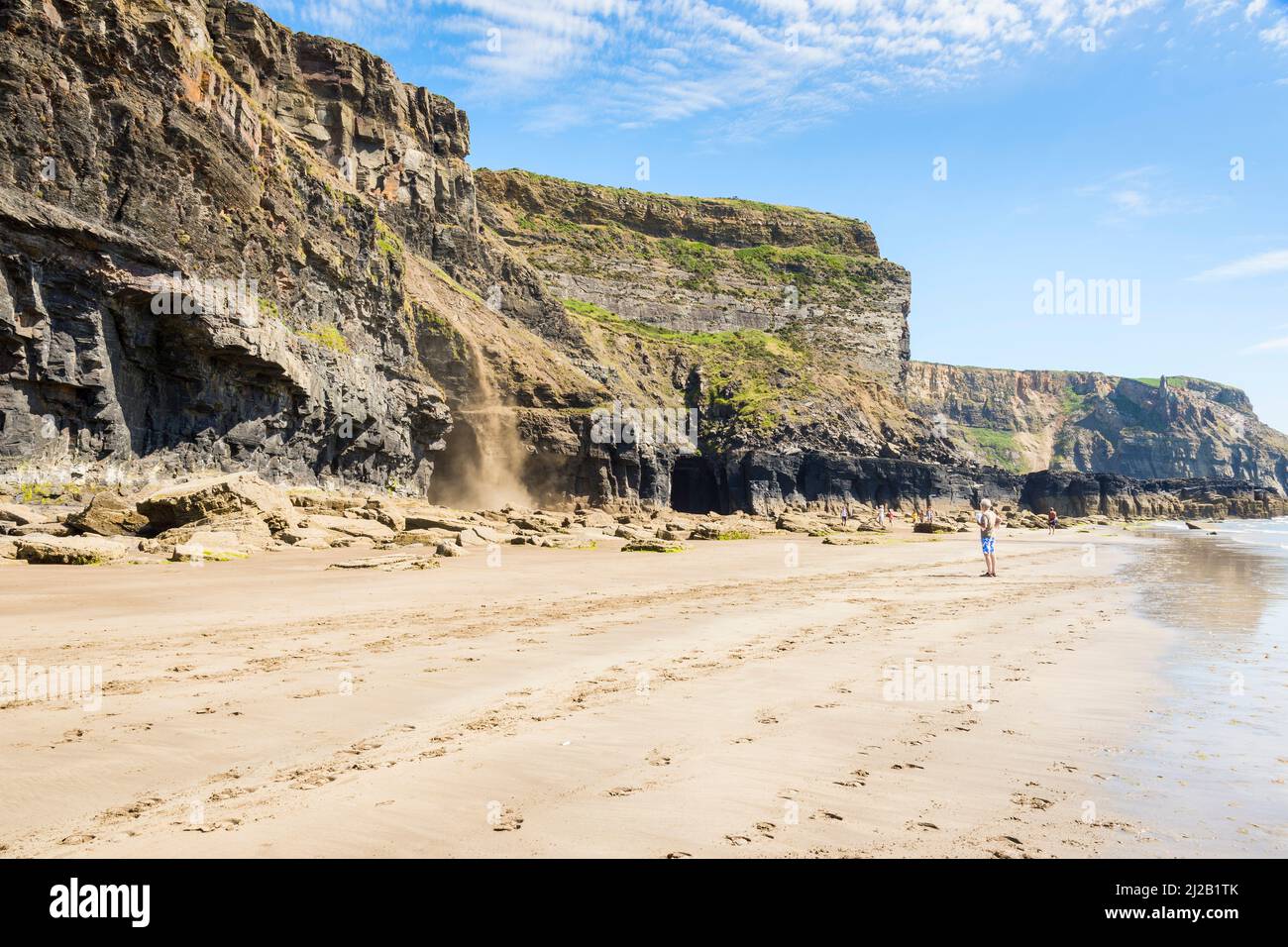 A man watches from the beach as rocks and dirt slide down the cliff face on a bright sunny day at Druidstone in Pembrokeshire, Wales. Stock Photo