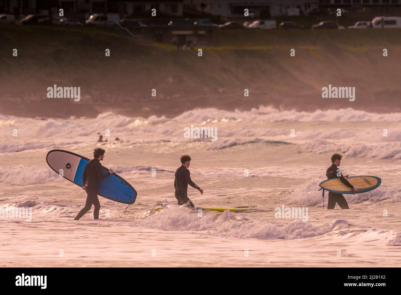 Surfers carrying their surfboards at the start of their surfing session in early evening light at Fistral in Newquay in Cornwall in the UK. Stock Photo