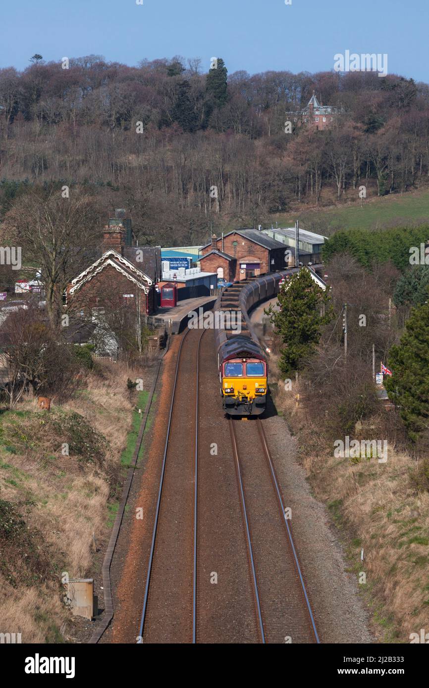 DB Schenker class 66 diesel locomotive 66143 in EWS livery passing Lazonby & Kirkoswald railway station with a merry go round coal train Stock Photo