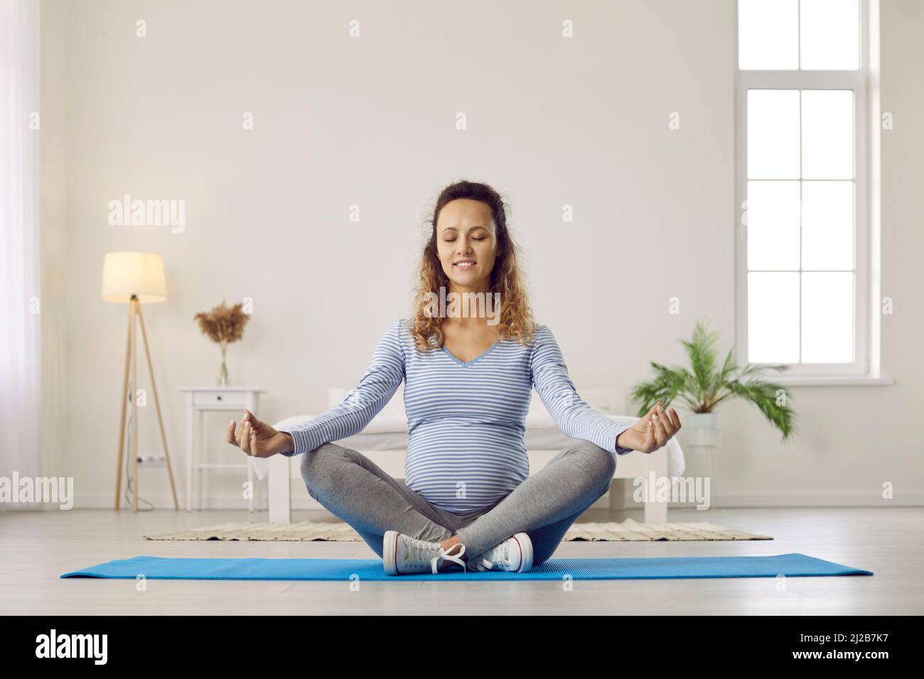 Calm pregnant woman meditates sitting in padmasana yoga position on floor at home. Stock Photo