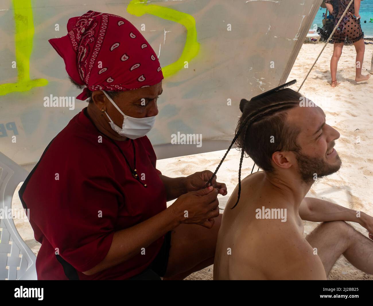 San Andres, Colombia - November 17 2021: Brown Woman with Mask Braiding a White Man's Hair Stock Photo