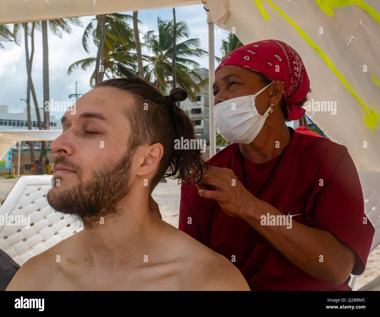 San Andres, Colombia - November 17 2021: Brown Woman with Mask Braiding a White Man's Hair Stock Photo