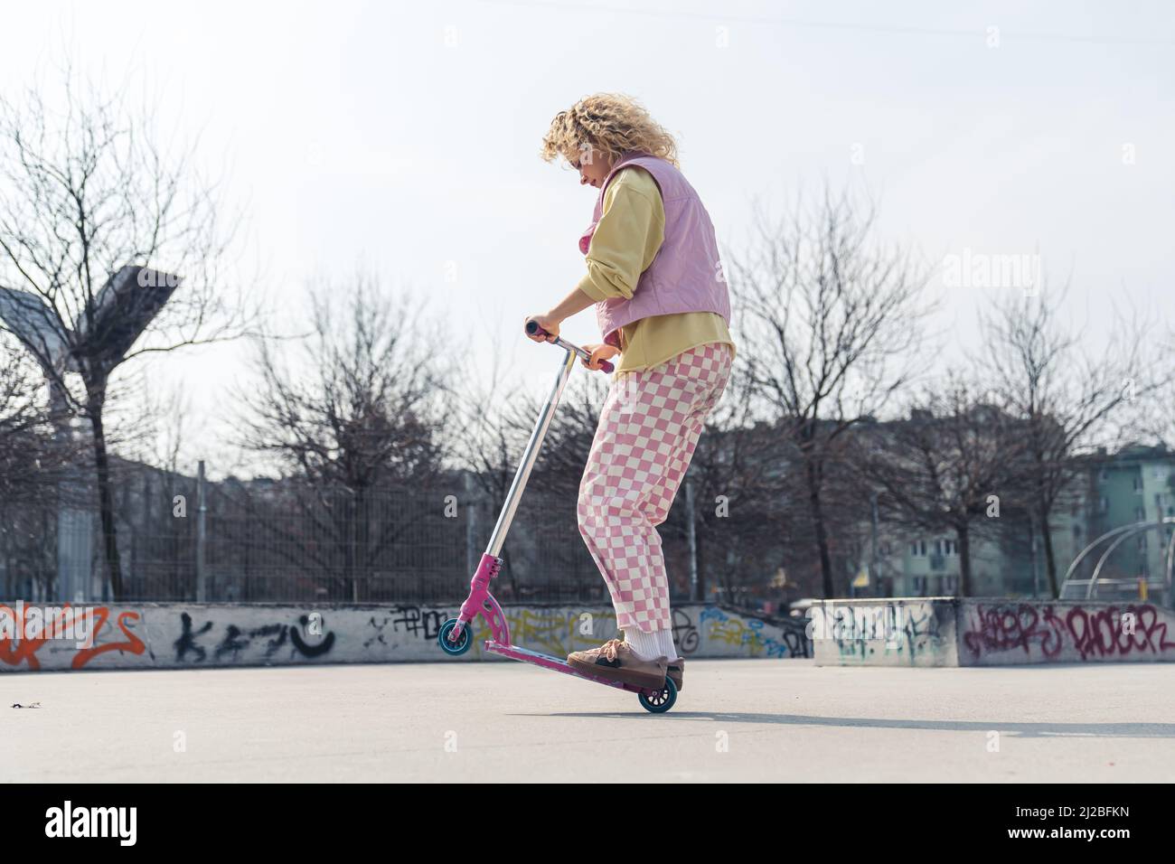 Pretty young blond curly haired woman drives a pink scooter on the skateboard ground casual attire copy space full shot . High quality photo Stock Photo