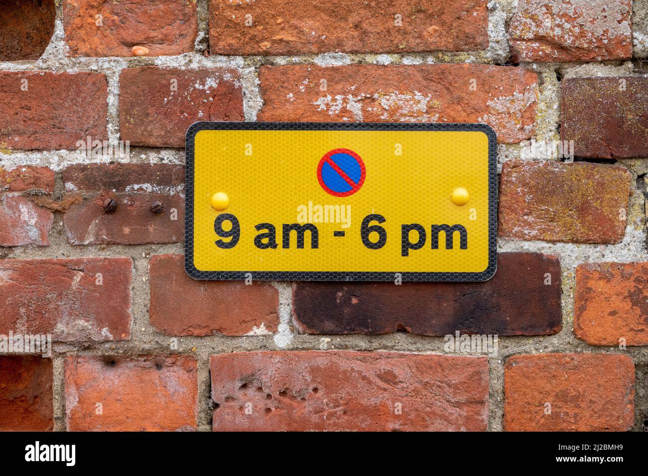 Small rectangular parking restriction sign black on yellow with red and blue symbol for the hours of 9am to 6pm mounted on a red brick wall Stock Photo