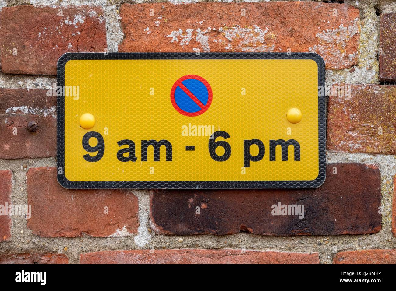 Small rectangular parking restriction sign black on yellow with red and blue symbol for the hours of 9am to 6pm mounted on a red brick wall Stock Photo