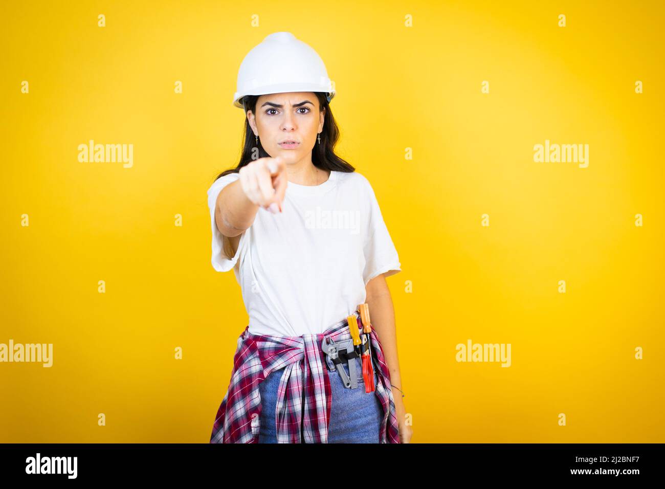 Young caucasian woman wearing hardhat and builder clothes over isolated yellow background pointing with finger to the camera and to you, confident ges Stock Photo