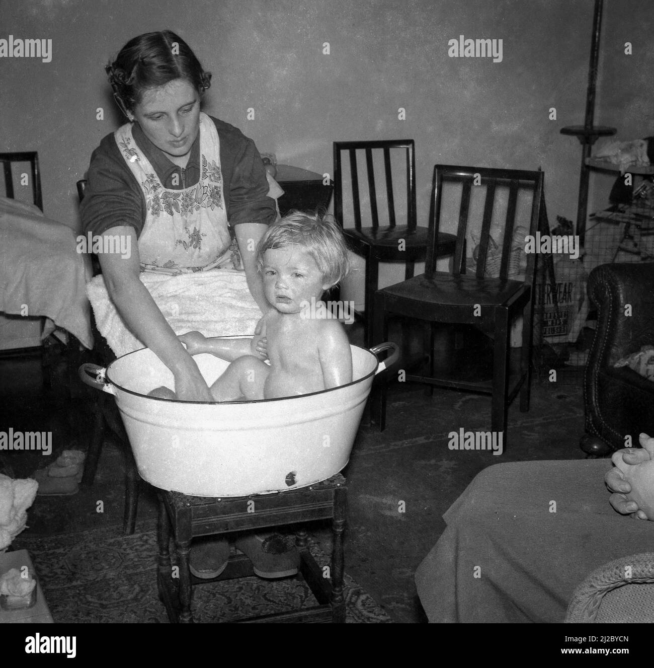 1950, historical, inside a front room, a mother in a pinafore giving her small child a wash in an enamel basin or bowl, which is sat on a foot stool, most probably positioned in front of the fire, Stockport, England, UK. Such bowls - made out of pressed steel with an enamel coating - were a common feature of households in this era, and as well as being used to wash clothes, were also used to give babies and infant children a bath, often sat infront of a coal fire. Stock Photo