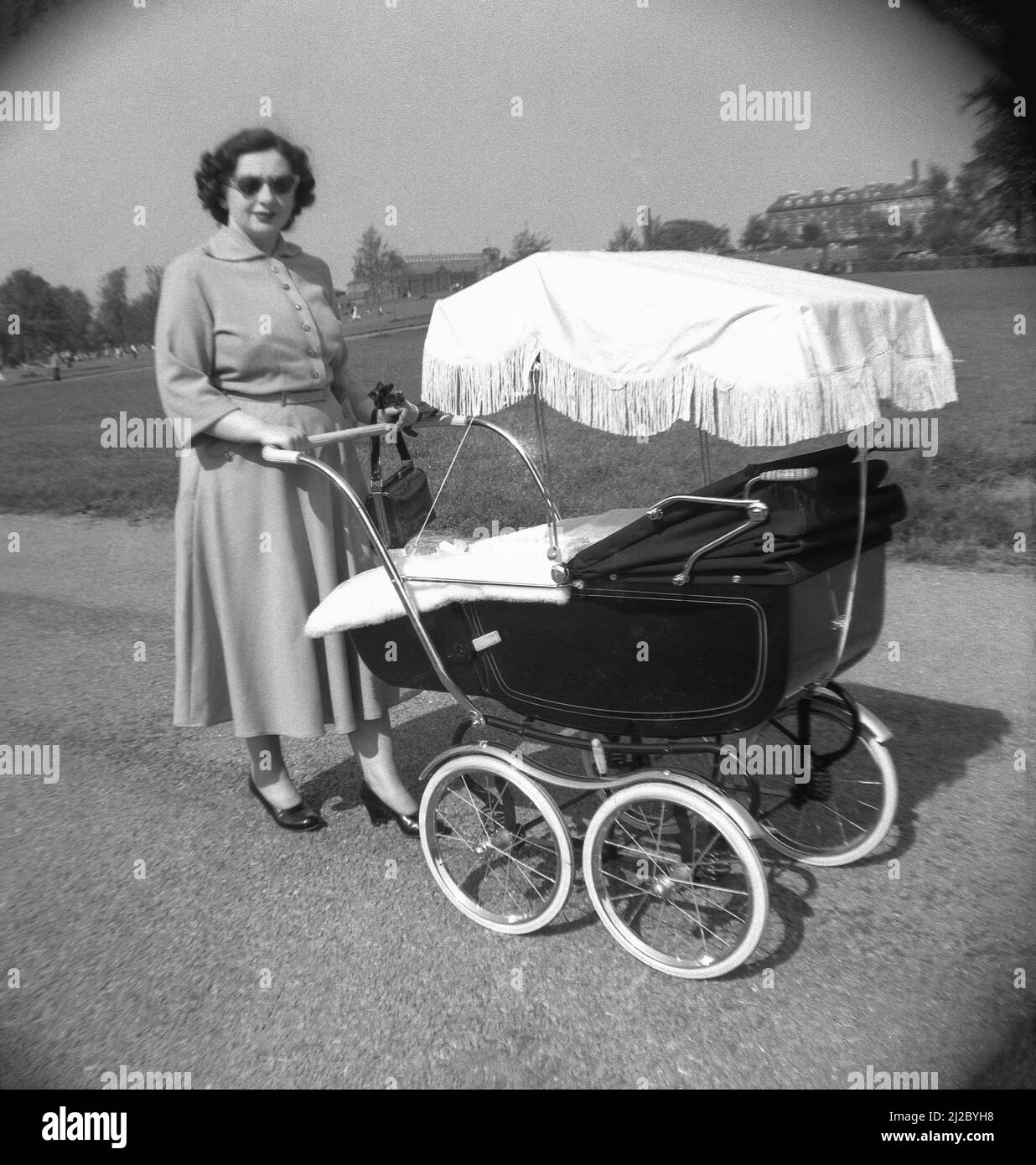1950s, historical, outside on a path in a park, a lady in a dress and shoes with a coach-built pram or baby carriage of the era, with shade, Shrewsbury Park, England, UK. A traditional coach-built pram was a popular item in this era, having many benefits for both mother and child. A sturdy steel chassis, suspension and large wire wheels meant it was light to push, while a padded mattress in a deep, solid pram meant a comfortable ride for the baby. An added advantage was the baby's position, high-up, facing the parent, meant plenty of eye contact. Stock Photo