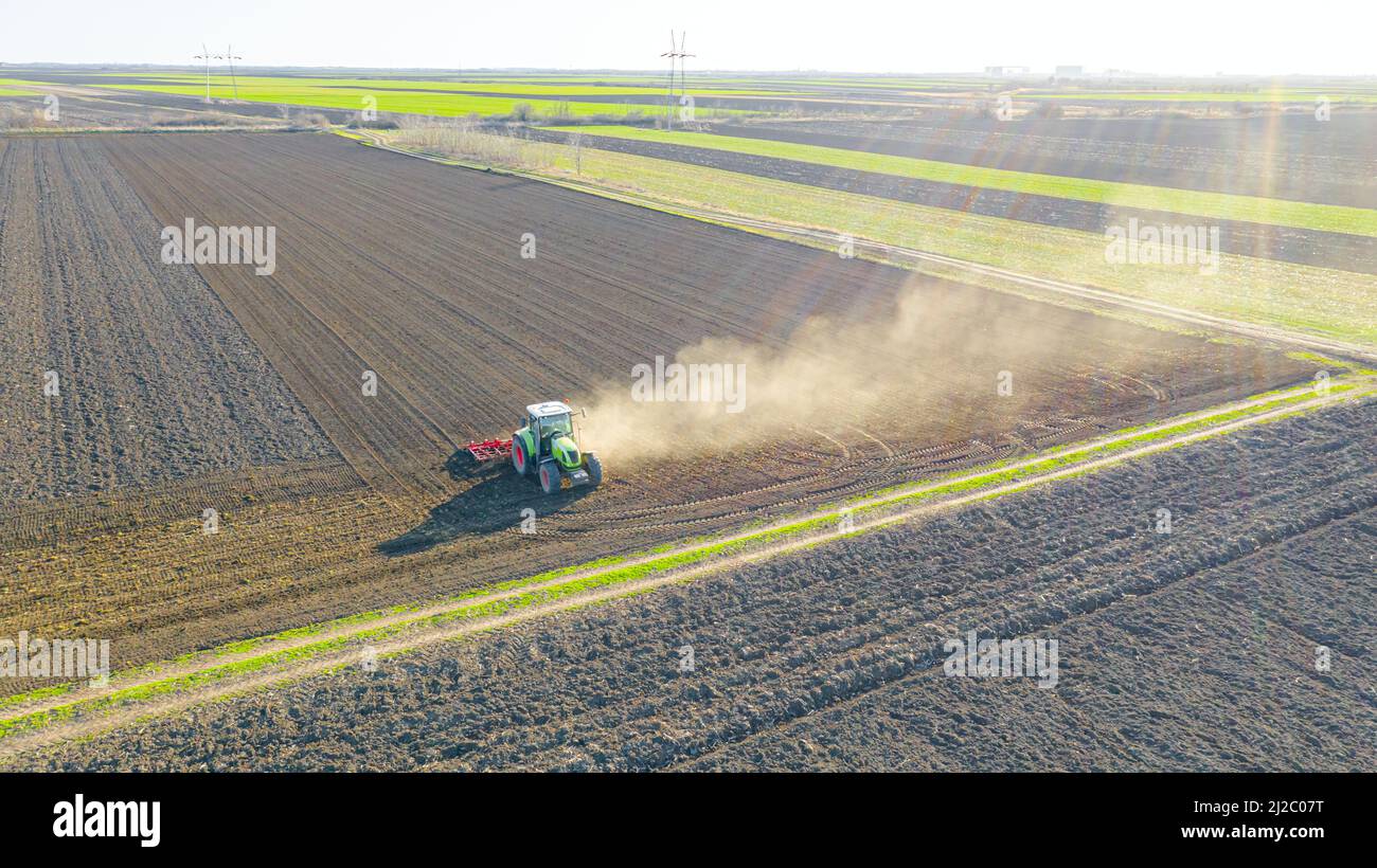 Above view of tractor as pulling machine, harrowing arable field, preparing soil for planting new crop next season. Cloud of dust is flying behind. Stock Photo