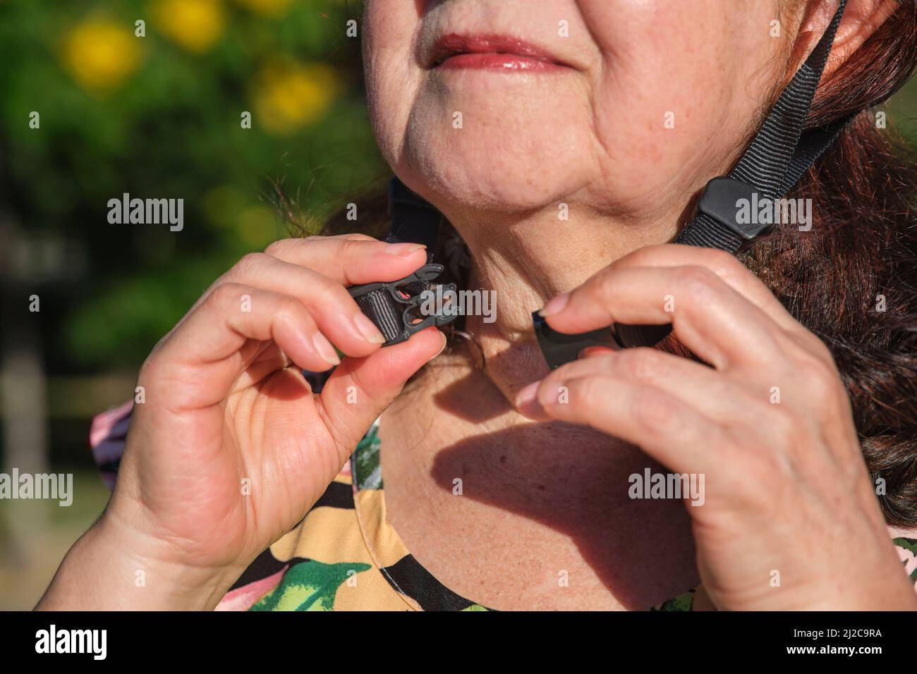 Mature hispanic woman putting on a sports helmet; close up image, detail of the hands fastening it. Concepts of active urban life and road safety. Stock Photo