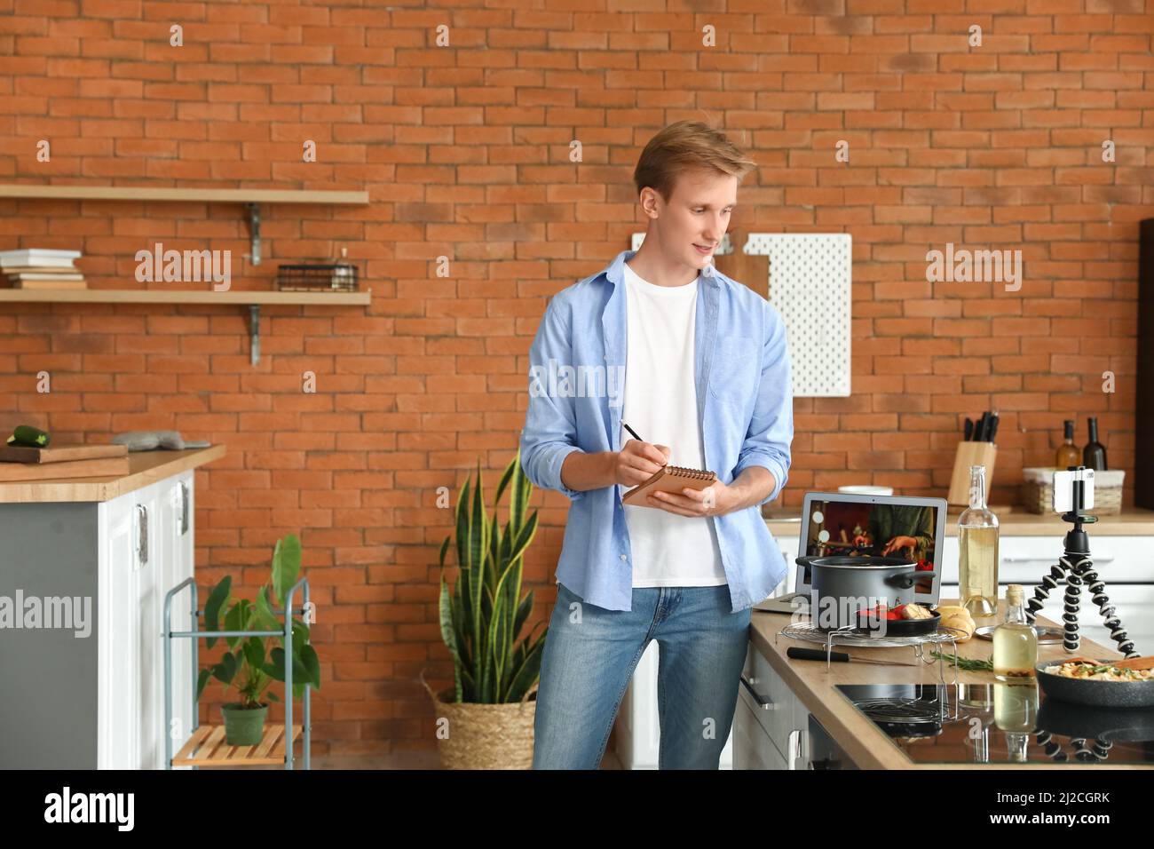 Young man making notes while watching video tutorial in kitchen Stock Photo