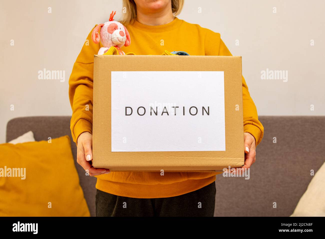 Young woman sitting on couch preparing parcel for sending to needy human. Girl with big kind heart puts used clothes new wear and soft bear toy in Stock Photo