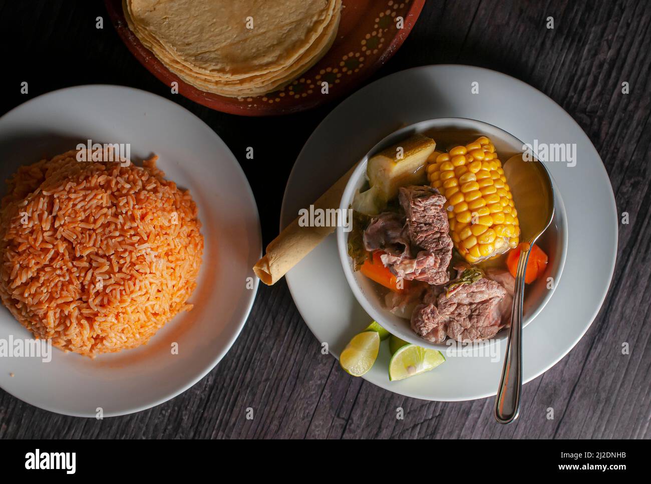 Mexican food, beef rib broth with vegetables on a white plate with slices of lemon and Mexican rice with tortillas on an old wooden table. Stock Photo