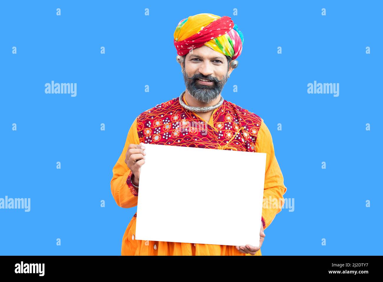 Portrait happy beard indian rajasthan man wearing traditional colorful outfit and turban holding blank board on blue studio background, advertising sh Stock Photo