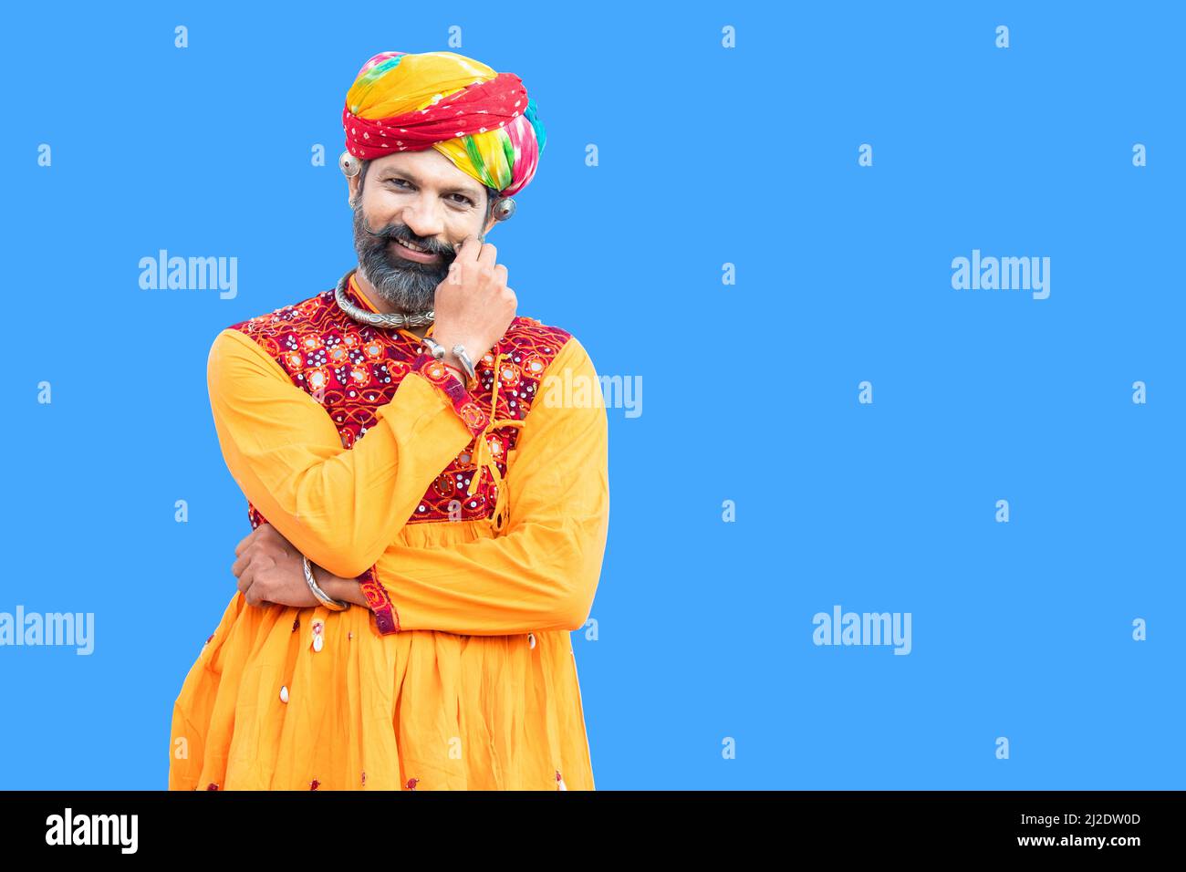 Portrait of happy beard indian rajasthan man wearing traditional colorful outfit and turban touch mustache pose against blue background. advertisement Stock Photo
