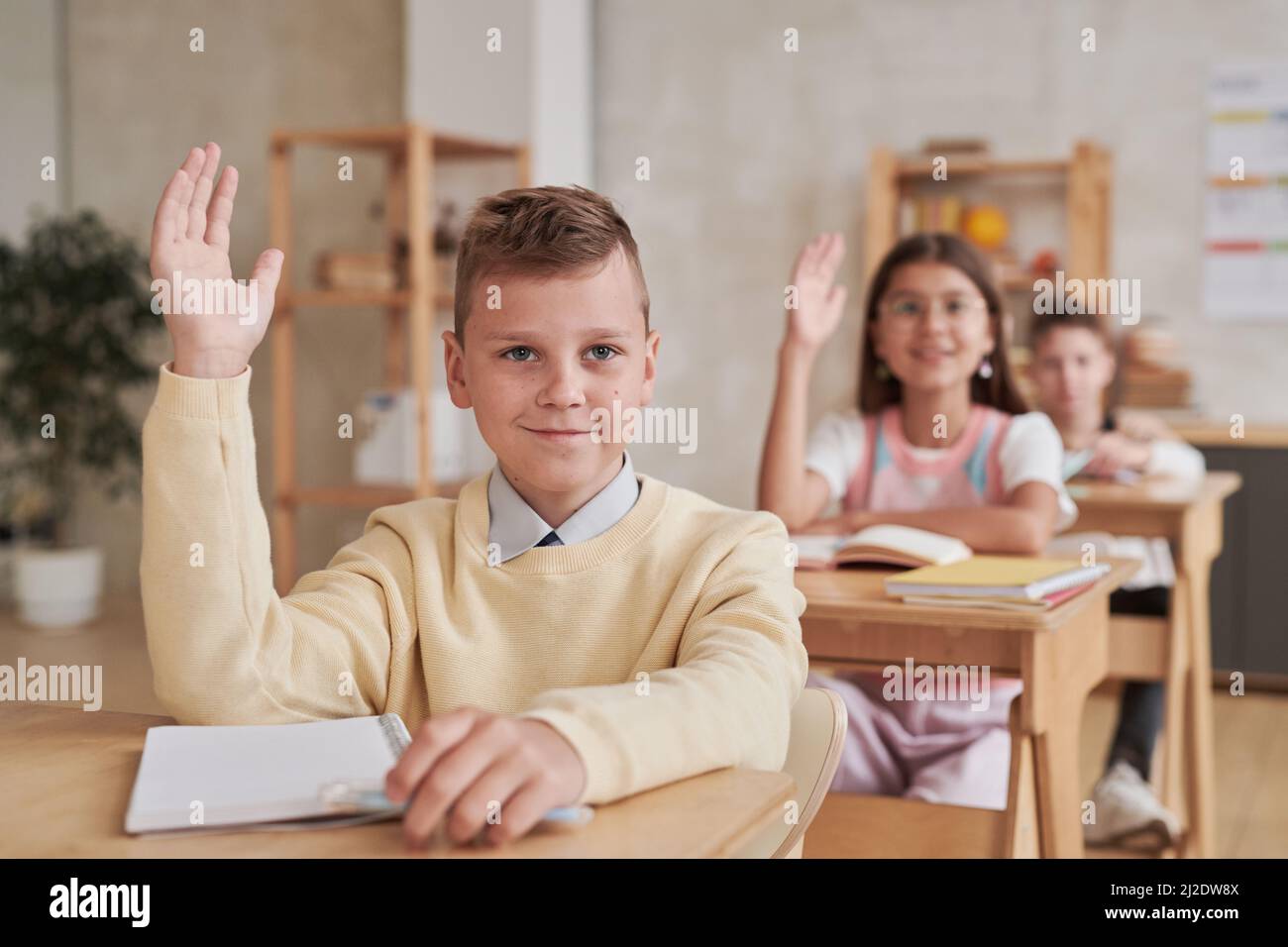 Portrait of smiling schoolboy raising hand while sitting at desk in ...