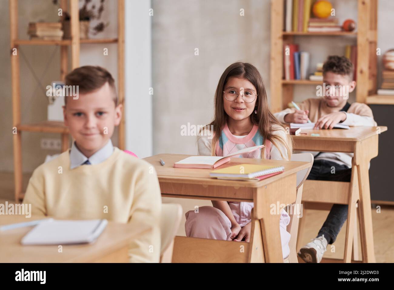 Portrait of cute schoolgirl wearing glasses and listening to teacher while sitting at desk in classroom, copy space Stock Photo