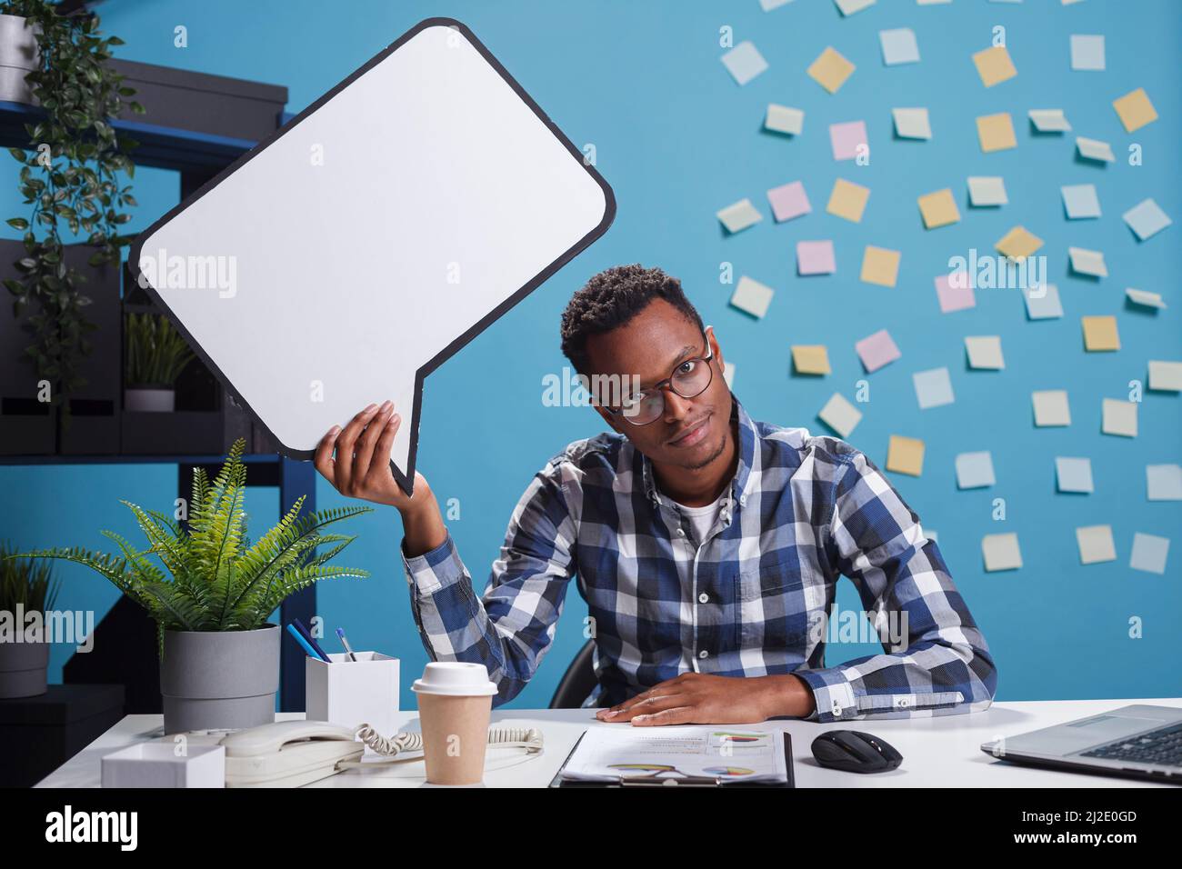 Bored office worker holding cardboard speech bubble sign above head while sitting in company workspace. Business development agency employee with speech bubble sign overhead. Stock Photo