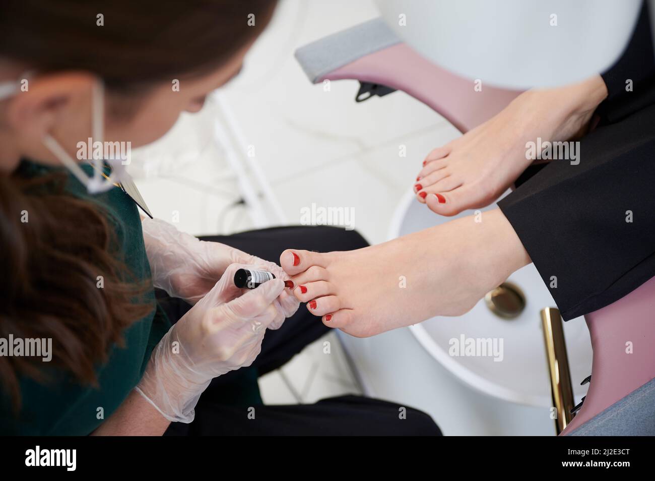 Nail specialist doing pedicure for client in beauty salon. Close up of female hands in sterile gloves touching woman foot while applying red nail polish on toenails. Stock Photo