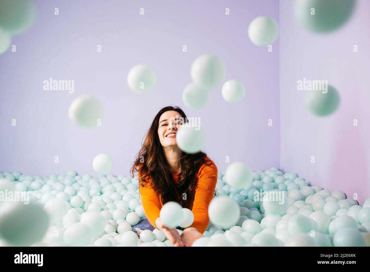 Portrait of brunette caucasian young woman with long hair, sitting in a swimming pool filled with balls and throwing them to the air Stock Photo