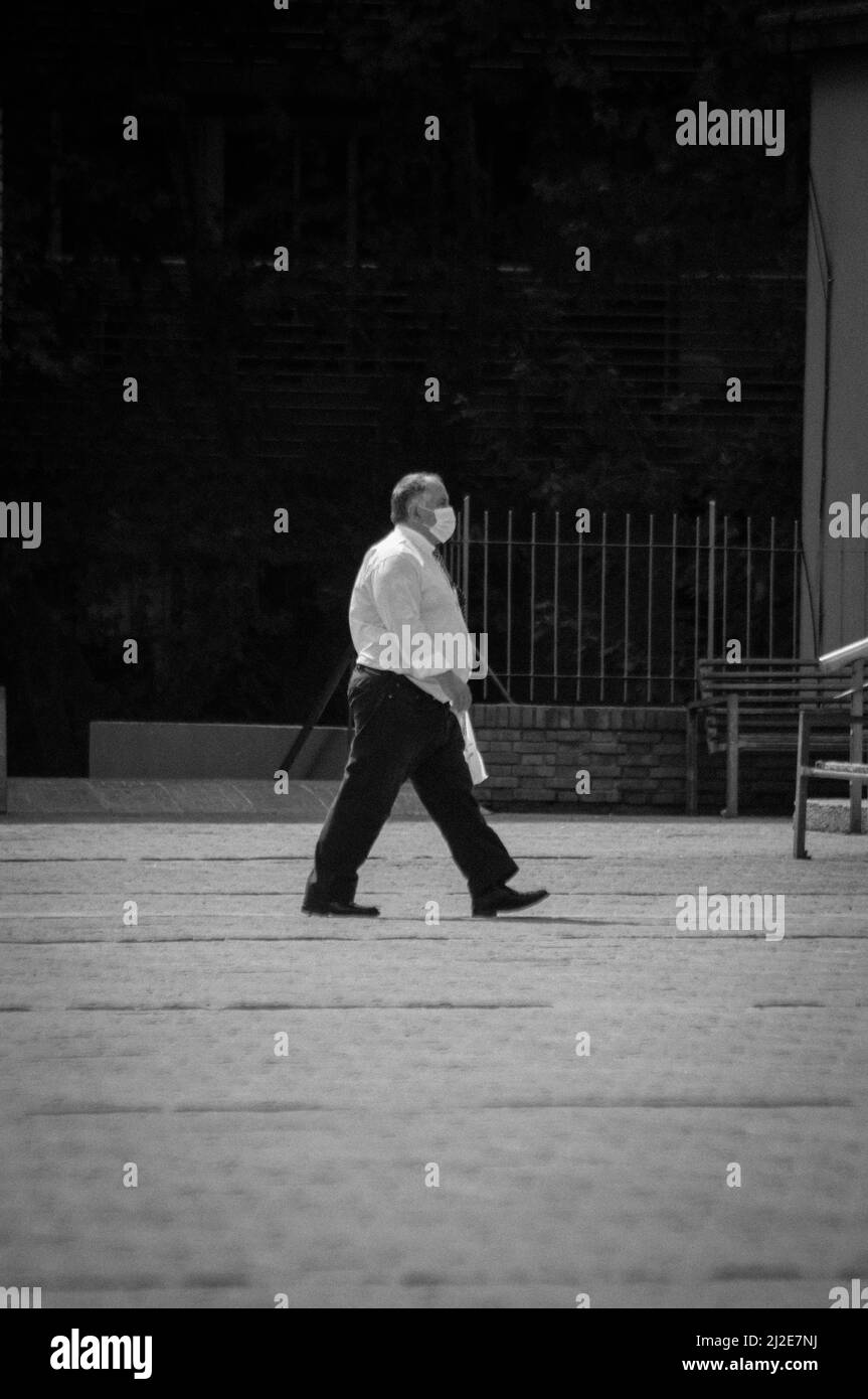 A vertical distant shot of an elderly male walking on the street with medical mask in black and white Stock Photo