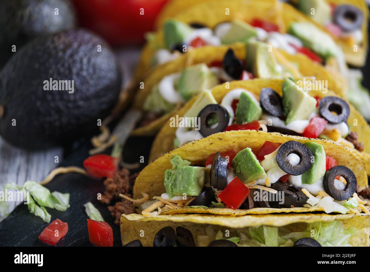 Tacos made of corn tortillas, ground beef, shredded cheddar cheese, black olives, avocados, lettuce, sour cream and tomatoes. Selective focus with blu Stock Photo