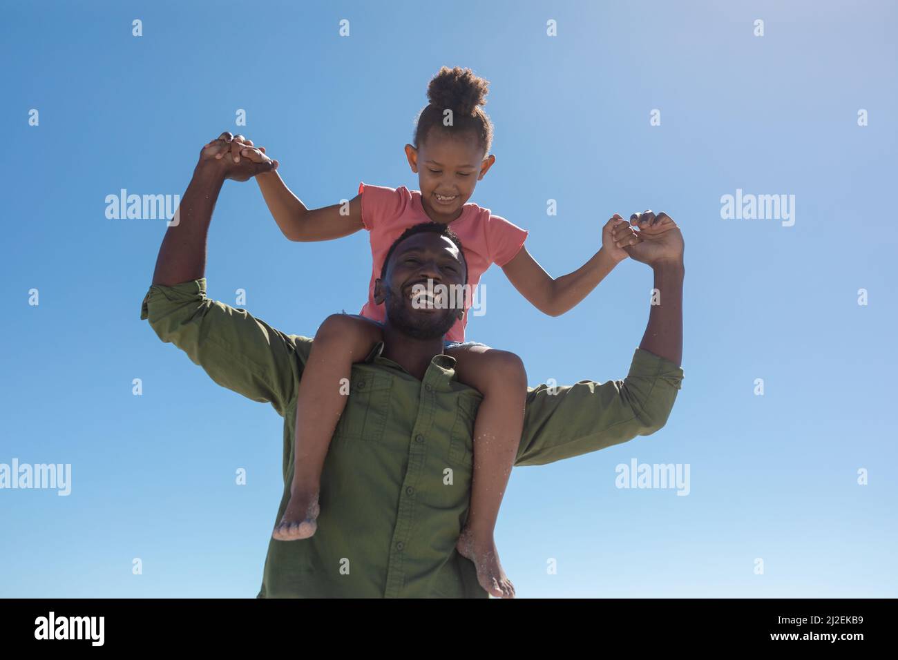 Low angle view of happy african american man with daughter sitting on his shoulders against blue sky Stock Photo