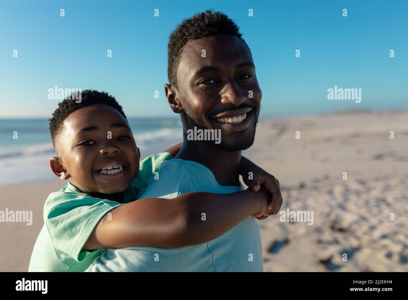Portrait of happy african american father piggybacking son at beach on sunny day Stock Photo