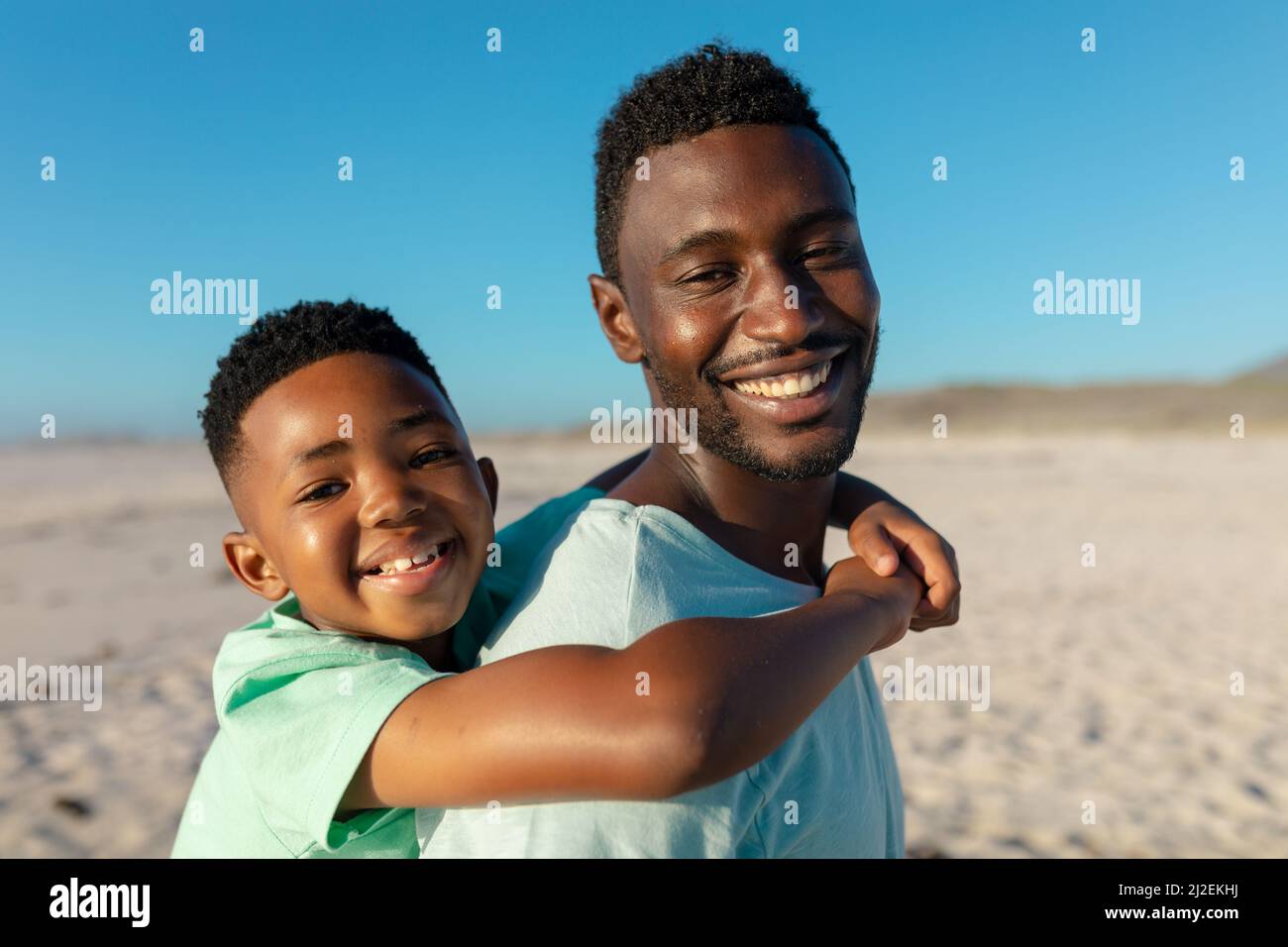 Side view of portrait of smiling african american father piggybacking son at beach on sunny day Stock Photo